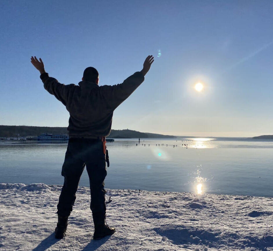 Energiearbeit und Freiheit in der Natur Berlin „Dennis Jahnke, Coach und energetischer Heiler, steht mit erhobenen Armen und gespreizten Beinen in X-Form am schneebedeckten Seeufer, Sonnenlicht spiegelt sich auf der Wasseroberfläche“